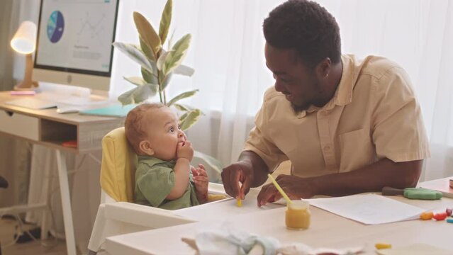 Caring African American father cleaning high chair after feeding his cute baby son at home