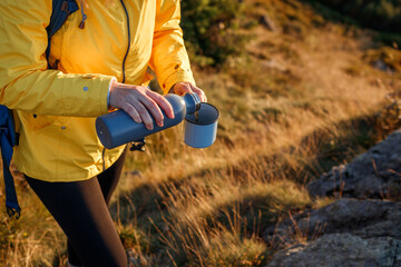 Hiker is pouring hot drink from thermos into travel mug in mountains