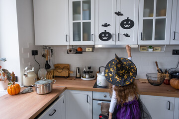 Child decorates the kitchen in home for Halloween. Girl in a witch costume plays with the decor for the holiday - bats, jack lantern, pumpkins. Autumn comfort in house, Scandi-style kitchen, loft