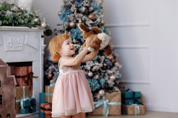 A child girl in a pink dress plays in a plush hare on the background of a decorated Christmas tree in a New Year's festive interior.