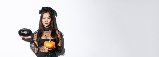 Sassy attractive asian girl in witch costume, holding pumpkins and looking at camera, standing over white background