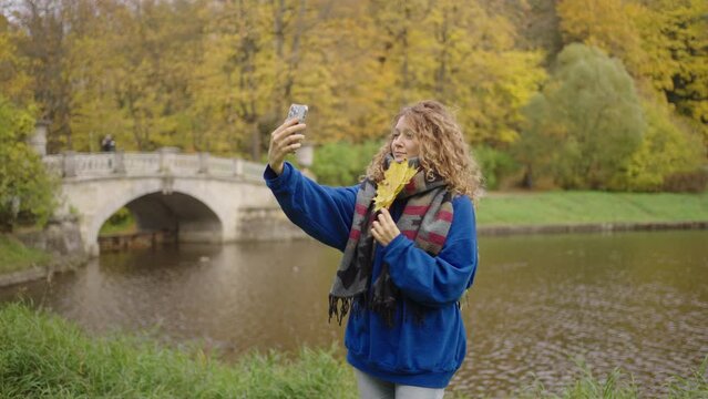 Beautiful woman takes a selfie on a smartphone in the park in a blue sweater with yellow maple leaves