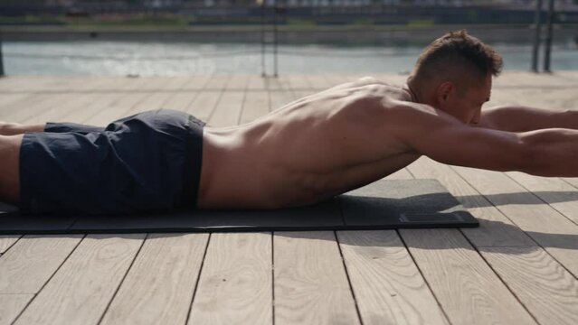 A fit caucasian man does Superman Back Extensions on a black mat. Isometric exercise. Working out and training lower back muscles outdoor.