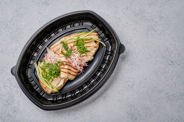 Bruschetta with smoked eel, young cheese and cucumber in black plastic container, top view. Designer food concept for take away or delivery