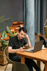 Man in a coffee shop sitting using cell phone and listening to music, on the table a computer and a cup of coffee.