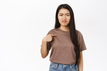 Obraz premium Sad asian woman pointing at herself, sulking and grimacing upset, being unconfident, standing over white background