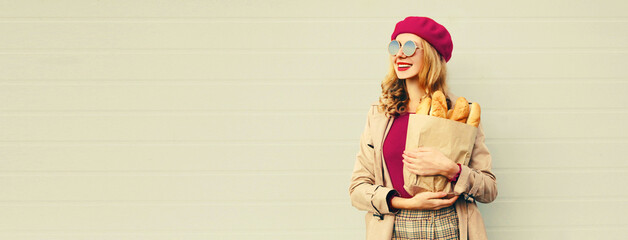 Portrait of happy smiling young woman holding grocery shopping paper bag with long white bread baguette on blank copy space gray background