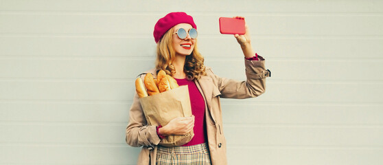 Portrait of stylish modern smiling woman taking selfie with phone holding grocery shopping paper bag with long white bread wearing beret on gray background
