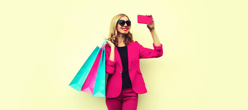 Portrait of beautiful smiling young woman taking selfie with smartphone holding shopping bags on background