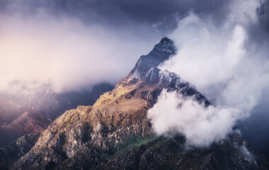 Mountain peak in low clouds at colorful sunset in Nepal. Dramatic landscape with beautiful high rocks in fog, moody sky, sunlight, tress, grass at sunset. Nature. Himalayan mountains. Scenery