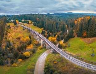 Fototapeta na wymiar Aerial view of railway bridge, railroad, rural dirt road, green meadows, trees, hills and cloudy sky in fall. Top view of beautiful old viaduct at sunset in carpathian mountains in autumn in Ukraine