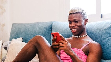 African Black transsexual man in stylish feminine clothes texting on cellphone on couch - obrazy, fototapety, plakaty