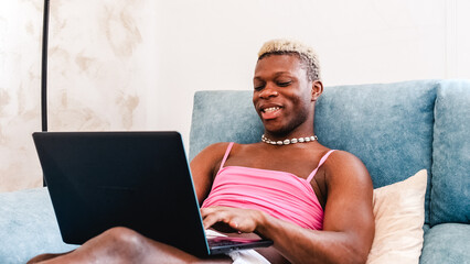 Transgender man using a laptop computer while relaxing sitting on a sofa at home - obrazy, fototapety, plakaty