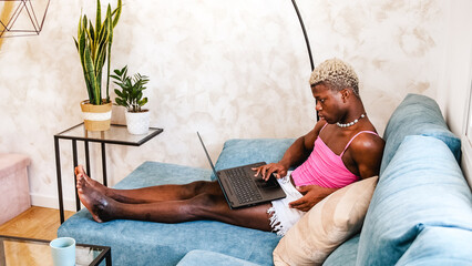 Serious transgender black African person using laptop in couch working at home