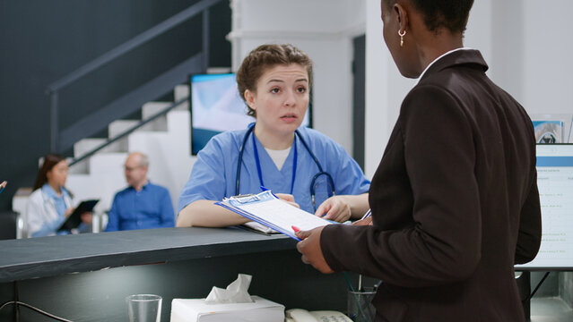 Diverse Medical Team Talking About Healthcare At Reception Desk, Working On Appointment Reports And Registration Forms. Nurse And Receptionist Giving Clinical Support In Facility Lobby.