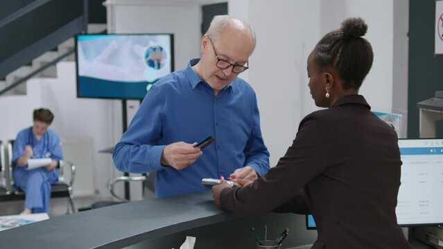Senior man paying with credit card at hospital reception desk, making electronic payment transaction for medical checkup appointment and treatment. Receptionist helping man to pay.