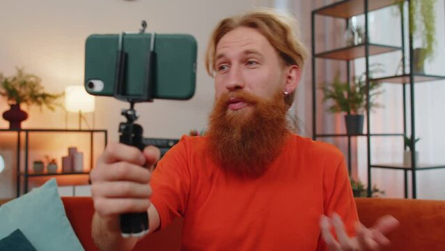 Portrait of caucasian bearded man blogger taking selfie on smartphone tripod, communicating video call online with subscribers. Young guy at home apartment in room sitting on orange couch. Lifestyle