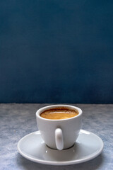 Cup of coffee with spoon on a white textured surface in front of a blue background. It has negative space at the top.