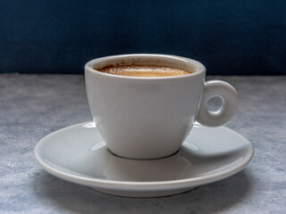 Cup of coffee with spoon on a white textured surface in front of a blue background. It has negative space at the top.