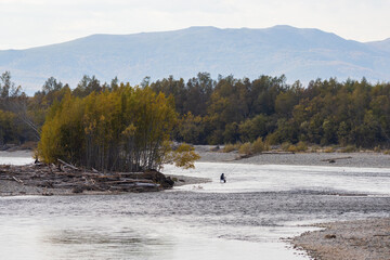 Beautiful autumn river landscape. A fisherman with a fishing rod sits on a chair and fishes in the river. Rest at nature. In the distance forest and mountains. Amateur fishing in the wild. Fall season