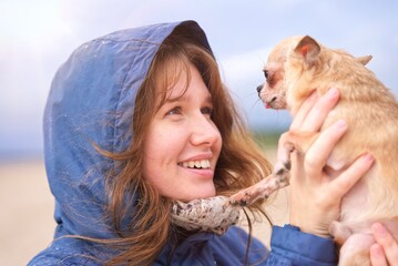 portrait of beautiful happy positive girl, young woman walking with her chihuahua or small dog, holding puppy on hands, smiling, having fun outdoors at beach
