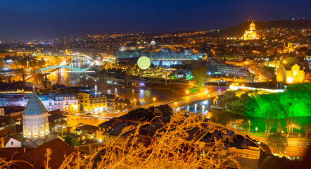 Scenic night view of Tbilisi Old town in colorful lights on banks of Kura river overlooking modern...