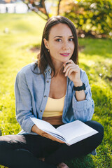 Fototapeta na wymiar Beautiful young caucasian woman holding an open book, reading book in park sitting on green grass. Healthy lifestyle, sport. Smart looking girl student. Summer