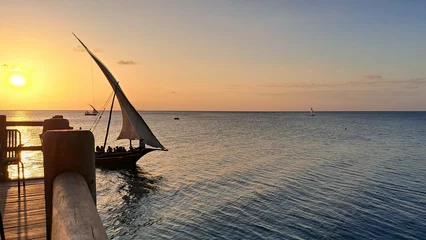 Photo sur Plexiglas Plage de Nungwi, Tanzanie Kendwa, île de Zanzibar, Tanzanie bateau boutre naviguant et une jetée de bungalow au toit de chaume en bois contre le soleil couchant et le ciel nuageux.