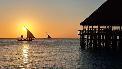 Photo sur Plexiglas Plage de Nungwi, Tanzanie Kendwa, île de Zanzibar, Tanzanie boutres naviguant et une jetée de bungalows au toit de chaume en bois contre le soleil couchant et le ciel nuageux.