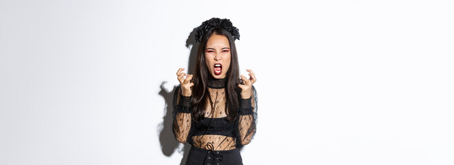 Image of mad woman in gothic lace dress and black wreath yelling angry and clenching fists, standing in halloween costume with furious expression, standing over white background