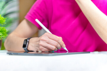 Female hand with wristwatch in bright crimson T-shirt sits at table with graphics tablet, holds stylus pencil in hands. Freelance illustrator, graphic designer, digital artist at work. Brainstorming 