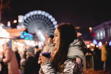 Young woman drinking mulled wine standing in Christmas market in city at night