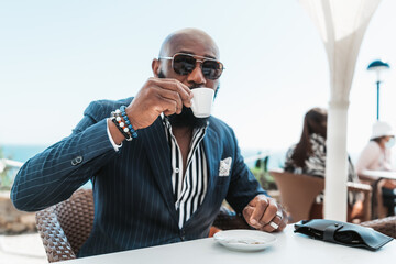 A portrait of a dapper bald African man in sunglasses and a fashionable summer suit sitting in a...