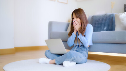 Young asian woman in good spirits working on laptop at home while sitting on the floor close to the couch. Excited female winner celebrating success while glancing at notebook