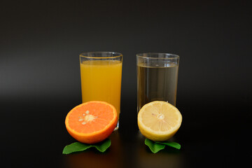 Two glasses with lemon and orange juice decorated with leaves and citrus halves on a black background.