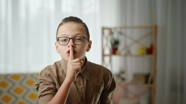 Boy asking to keep silence. Smart boy in brown shirt and stylish glasses looking around then touching lips and asking to be silent in living room at home.