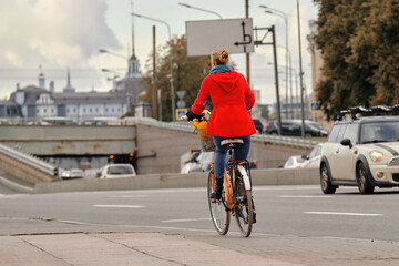 Obraz premium A girl in a red coat rides an orange bicycle around the city next to the auto road.
