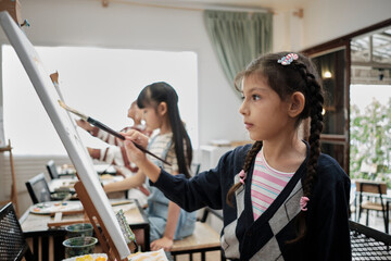 A little girl concentrates on acrylic color picture painting on canvas with multiracial kids in an...