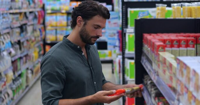 Caucasian young man buyer shopping groceries in supermarket, taking spaghetti from shelf, standing with shop cart indoors. Guy buys grocery, choosing food in super market