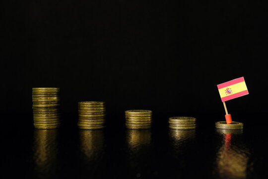 Spain economic recession, financial crisis and currency depreciation concept. Spanish flag in decreasing stack of coins in dark black background. - Powered by Adobe