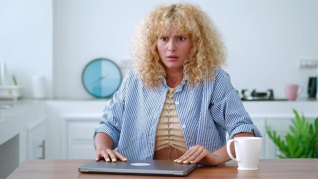 Anxious Caucasian young blonde woman, sitting at a wooden table at home kitchen, carefully reading information on a laptop monitor and suddenly expressing fright and numbness from what she has read