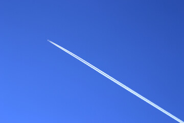 The contrail of the aircraft on the background of a clear blue sky. A distant passenger jet is flying at a high altitude in the blue sky and leaving a long white smoke trail behind it.