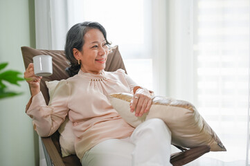 Asian aged-woman sipping coffee, laying on armchair, relaxing in her living room.