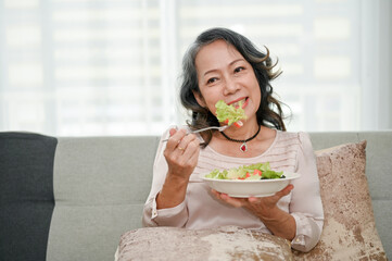 Wellbeing 60s retired Asian woman eating healthy salad while relaxing in her living room