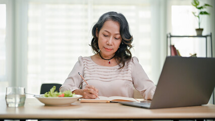 Beautiful senior asian businesswoman working from home, focused on writing at her desk.