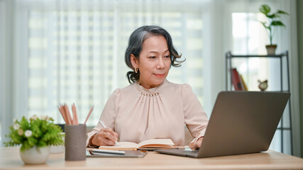 Charming 60s senior Asian female writer working at her desk, writing something on her book