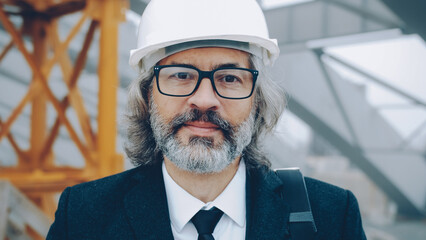 portrait of building project manager standing outdoors wearing helmet and suit looking at camera. Construction and senior specialists concept.