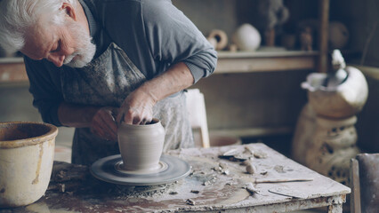 Fototapeta na wymiar Experienced ceramist grey-haired bearded man is smoothing molded ceramic pot with wet sponge. Spinning throwing wheel, muddy work table and handmade clayware are visible.