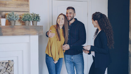 Real estate agent is meeting happy married couple, showing papers and telling them about new apartment. Young people are excited, they are looking around and hugging. - 537420691