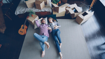 Top view of loving young couple lying on bedroom floor in new apartment and talking. Guitar, packed...